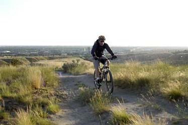 Man on mountain bike rides a dirt trail with green grass on each side and broad cityscape in background