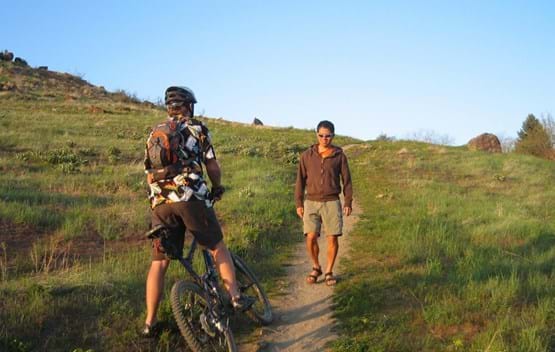 Hiker walking on dirt trail. Mountain biker pulled to the side of the trail and stopped to allow hiker to pass. Green grass on both sides with blue sky.