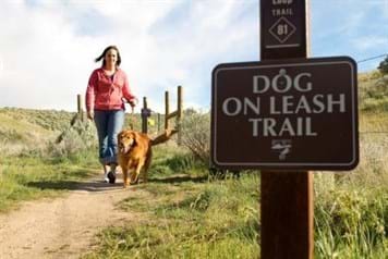 Woman walks on trail with leashed dog. Large sign in foreground reads "Dog On Leash Trail".