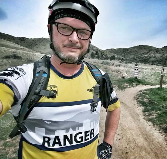 Man with bike helmet standing on trail with yellow and white jersey that says "Ranger".